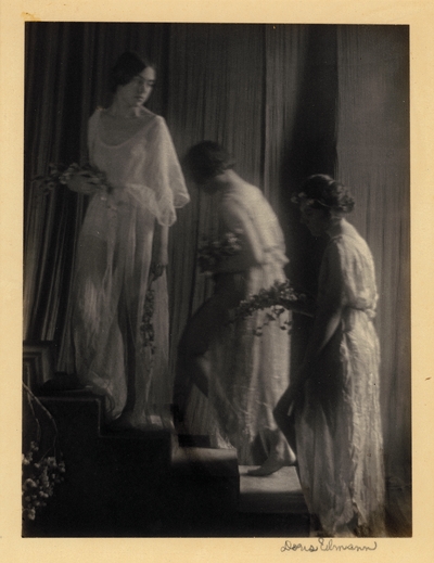 Three dancers in classical garb, holding flowers, ascending steps with curtain in background