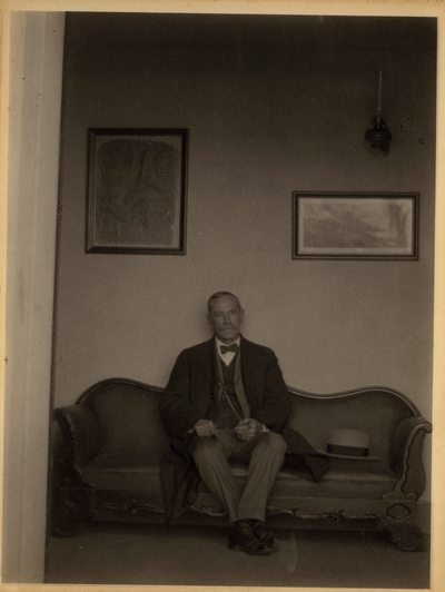Elderly man with mustache, in suit and bow tie, sitting on sofa with hat beside him and pictures hanging on the wall