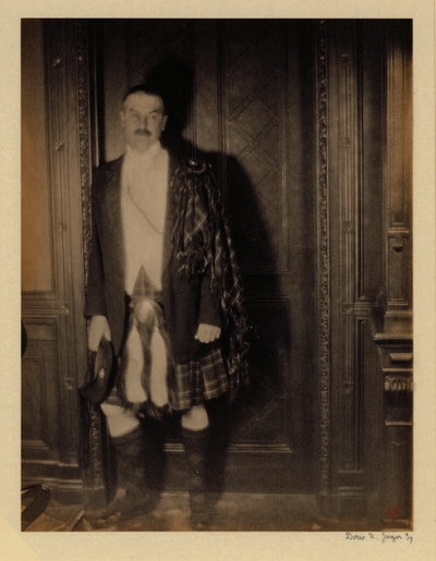 Man with mustache, in Scottish garb with kilt, standing in front of wood paneled doorway