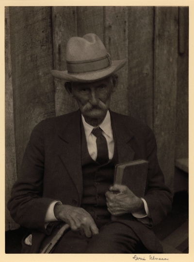 Elderly man in hat, mustache, suit, and tie, seated with book and cane