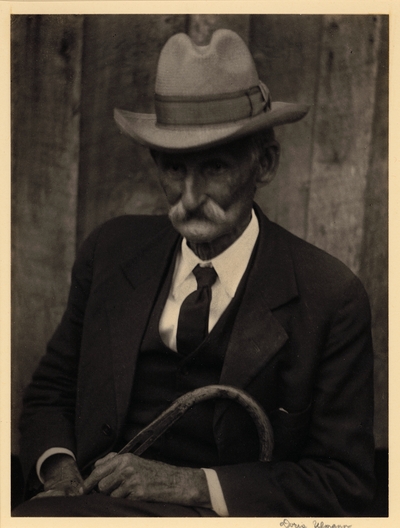 Elderly man with mustache, in hat, suit, and tie, seated with cane