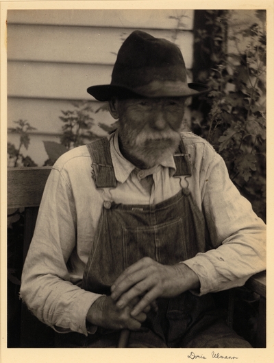 Elderly, bearded man in hat and overalls, seated with cane