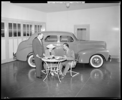 Lexington Nash Motors, 152 Rose; interior of car dealership, car                             parked inside; a woman and man are seated at a table in front of                             vehicle, man standing next to woman while flipping through a                             brochure