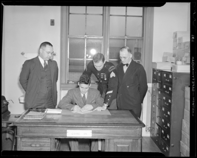 United States Marine Corps, recruiting office; group of men                             standing next to a desk as a young man signs paperwork