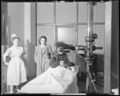 Cancer Ward; Good Samaritan Hospital, 310-330 South Limestone;                             patient (child) lying on table under apparatus (cancer treatment);                             nurse, man, and woman standing beside child