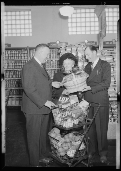 Kroger Store; 726 East Main; interior; woman standing beside her                             shopping basket, two men presenting woman a basket of grocery                             coupons