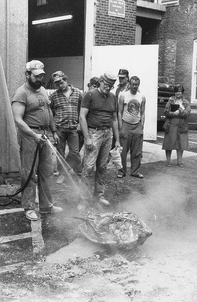 Jack Gron, John Tuska and several unidentified people standing outside the University of Kentucky foundry. Gron is spraying water from a hose on the John Sherman Cooper bust mold to remove the plaster