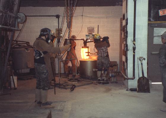 Scott Oberlink, Jack Gron, an unidentified man and Andrew Marsh lifting a crucible out of the furnace in the University of Kentucky foundry for the casting of 