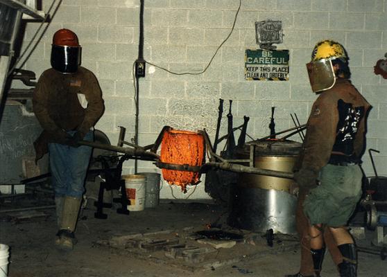 An unidentified man and Andrew Marsh lifting a crucible out of the furnace at the University of Kentucky foundry for the casting of 