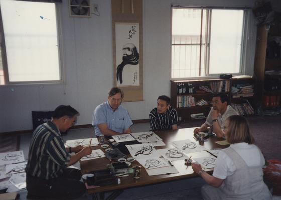 An image a Japanese man named Ohsawa-san, a daruma painter, painting on a table with Irwin Pickett, Heike Pickett, and two unidentified Japanese men watching. John Tuska visited Japan for the Kentucky Arts Council