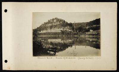 Multiple long low buildings on far bank, two large buildings with chimneys, two boats in foreground on near bank, notation                          Boone's Knob mouth of Hickman (Camp Nelson) 1907