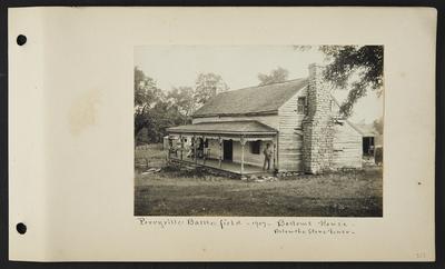House with stone chimney, one man standing on nearest edge of porch, one man leaning back in his chair and two women sitting at opposite end of porch, wooden fence extending from porch to small building behind house, notation                          Perryville Battlefield 1907 Bottom's House, below the stone fence