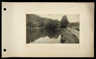 Large pond surrounded by trees, two white ducks swimming, two women and two children walking along road on far right