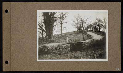 Wooden bridge running over dry creekbed, dirt road shored up with stones, wooden fencing along road and in background