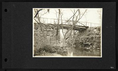 High wooden bridge with railing built over low river, road shored up with stones