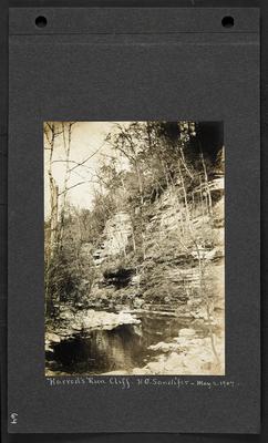 Creek with bluffs rising up the right hand bank, man standing on rocks on edge of creek with camera on tripod, notation                          Harrod's Run Cliff, H.G. Candifer, May 2, 1907
