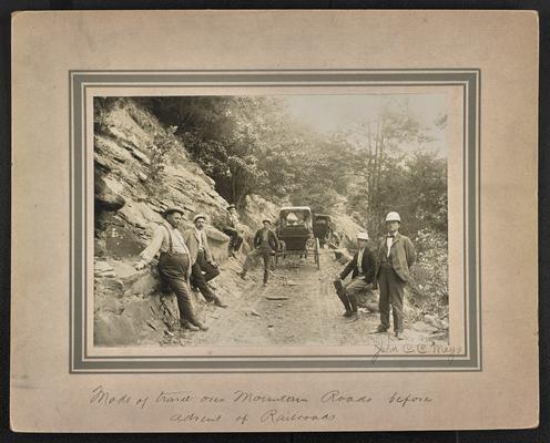 6 unidentified males resting on either side of a dirty road, two carriages stopped ahead of them. Back of the photograph reads: Mode of travel over Mountain roads before advent of railroads