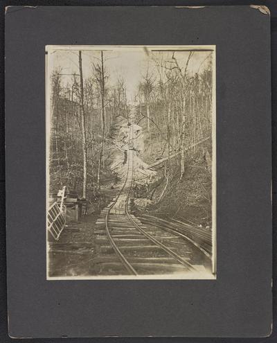 A railroad being built through a forest on a hill, two unidentified males pulling a cart on the tracks in the distance