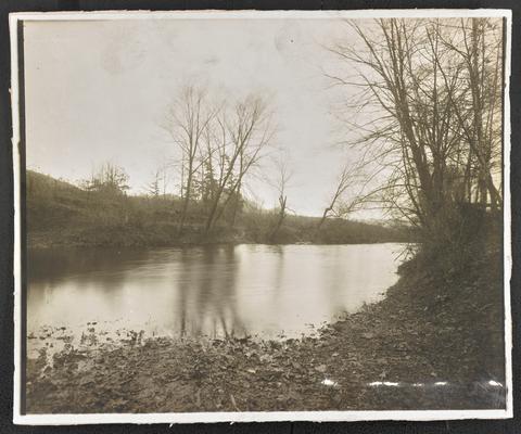 A pond surrounded by leaves and tress. The back of the photograph reads: The Mountain Child- His environment