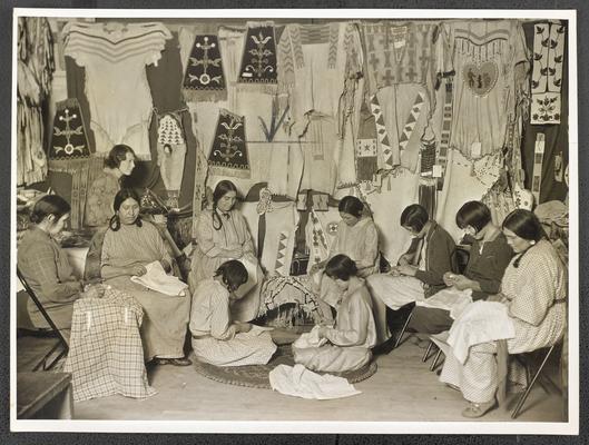 A group of women sitting sewing and sitting around displays of traditional Native American clothing