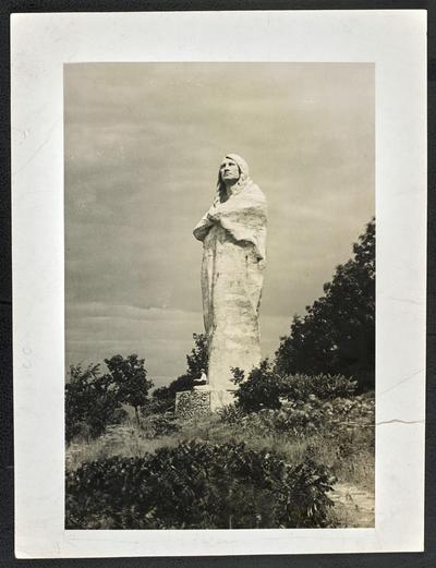 Unidentified man sitting at the base of an monument of a Native American male. The back of the photograph reads: Blackhawk Indian Memorial at Oregon, Illinois