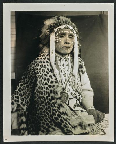 Formal portrait of a man wearing traditional Native American dress, including a headdress and facepaint. The back of the photograph identifies him as: Chief Wades in the Water