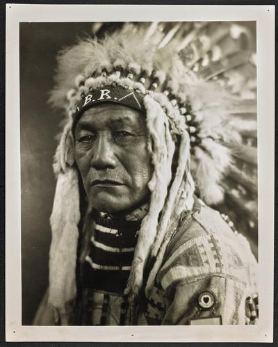 Formal portrait of a man wearing traditional Native American dress. Back of the photograph identifies him as: Chief Bird Rattler, a Judge on the Blackfoot Reservation