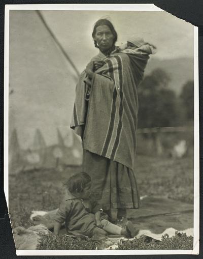 A woman with a baby on her back and a toddler at her feet, a teepee stands in the background. The back of the photograph identifies her as: Mrs. Eagle Calf, Blackfoot Reservation