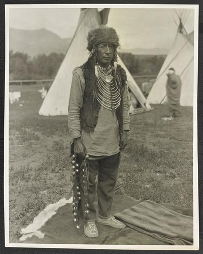 Full length portrait of a man wearing tradition Native American dress, standing in front of a teepee. The back of the photograph identifies him as: Chief Eagle Calf. Graduate of Carlisle, worked in newspaper depart. at Carlisle