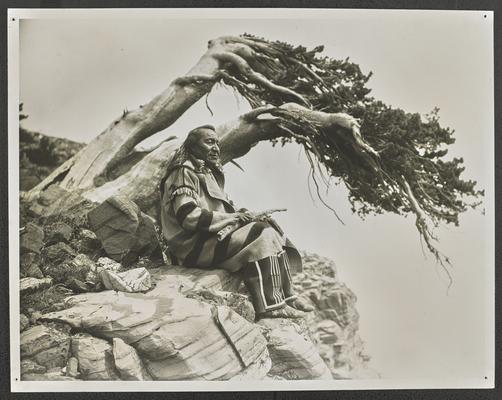 A man in traditional Native American dress, sitting at the edge of a cliff. Typed label identifies him as: Arrow Top Knot, aged 64. A distinguished member of the Blackfeet Tribe who made a fine record