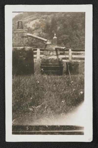 A moonshine still. The back of the photograph reads: The men that made this still are in Mr. Sizemore's moonlight school. They have rather peculiar names. Bill Couch (First Beard), John Sizemore (Coal Foot). They made lots of moonshine with this still