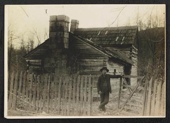 Unidentified man standing in front of a log cabin