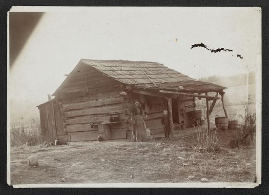 Unidentified woman, boy, and dog standing in front of a log cabin