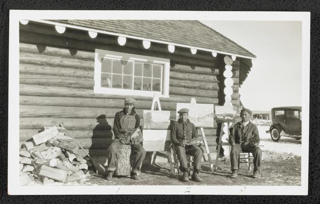 Three males sitting in front of paintings they have painted. Back of the photograph identifies them as: 1. Sam Calfrobe 2. Al Evans 3. Tom Dogtaking Gun