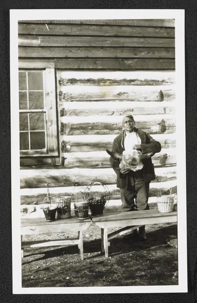 A man holding an animal skull and standing behind display tables with woven baskets. Back of the photograph identifies him as: Swims Under