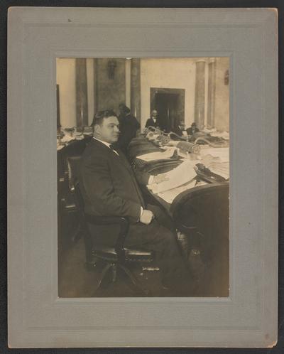 Male, unidentified, sitting in a suit at a desk. Back of the photograph reads: To Mr. and Mrs. E.D. McGuire, the picture on the reverse is the picture of the vandidate you helped to elect together with the chair to which he was elected. He sends you this picture merely to manifest in a small way his great love for you both not because of this but because you are just Boone and Stella. Your Brother, Bun