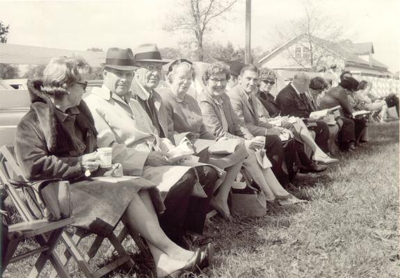 Groups; Unidentified; A group of people seated next to a fence