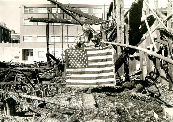 Ashland Bulk Oil Plant; 1973 Explosion and Fire; A man holds a frayed American Flag; burned building in the background