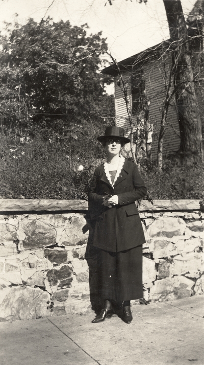 A portrait of an unidentified woman standing on a sidewalk next to a stone wall. This print was taken by De Luxe Studio, Hot Springs, Arkansas. This print was found pasted to the front of page 109 of 