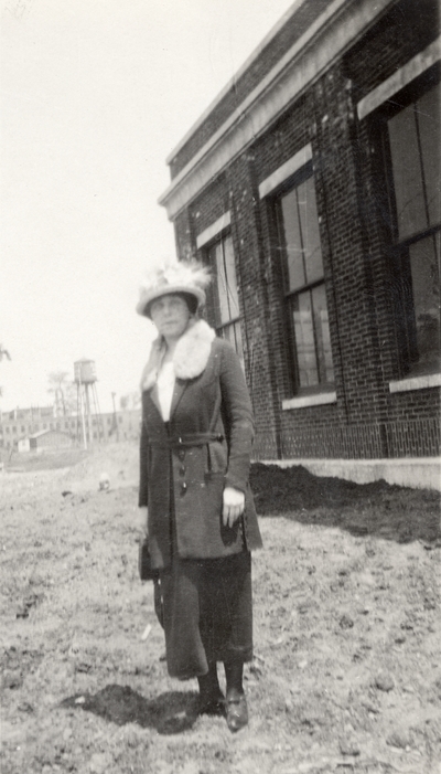 A portrait of an unidentified woman standing in dirt next to a building. This print was found pasted to the front of page 112 of 