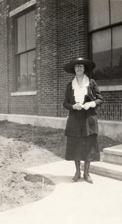 A portrait of an unidentified woman standing on a sidewalk next to a building. This print was found pasted to the front of page 112 of 