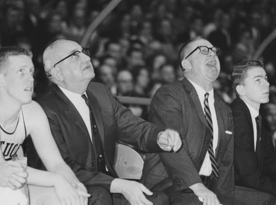 Basketball game action; photo of coaches Adolph Rupp (left) and Harry Lancaster from the bench; player Don Rolfes sits far left; man to the far right is unidentified