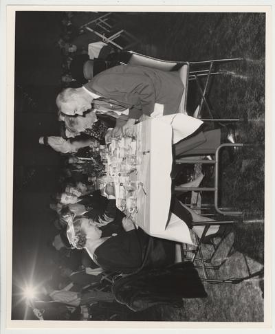 President Frank Dickey (left, facing the camera) is sitting at a table with his family.  Mildred (spouse of Governor Chandler) is the first person on the left, Governor A. B. 