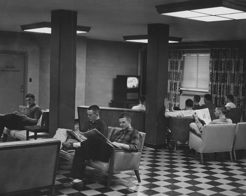 Five unidentified men are watching television and four unidentified men are reading newspapers in the basement of Donovan Hall