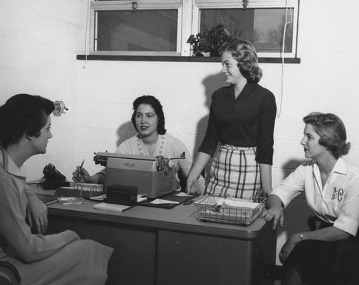 Four unidentified women are seated around a desk in Holmes Hall. Photographer: University of Kentucky. Received November 18, 1958 from Public Relations