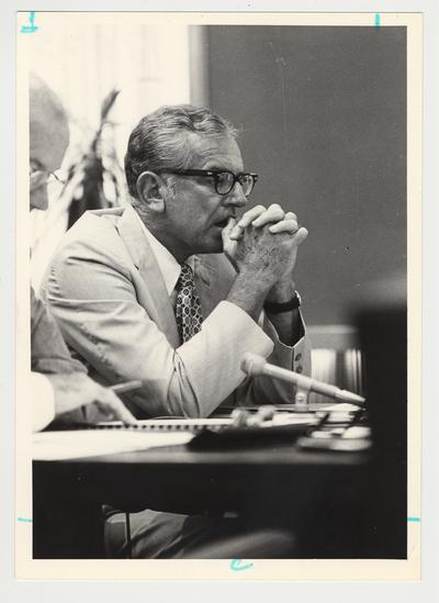 President Singletary is sitting at a table with his hands clasped in front of his face.  He is at an unidentified meeting