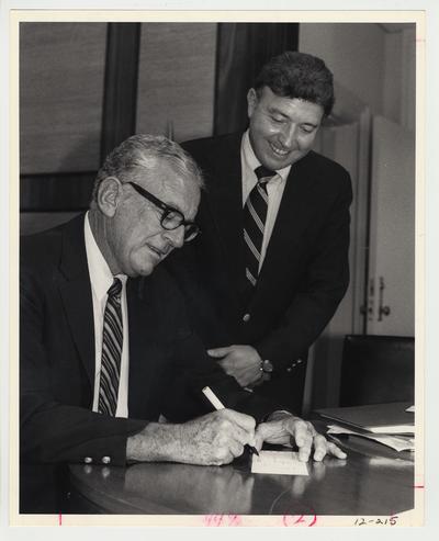 President Singletary (seated, left) is signing a piece of paper while John Bryant (standing, right), Assistant Director of the Library Department and head of the United Way drive at the University of Kentucky, watches