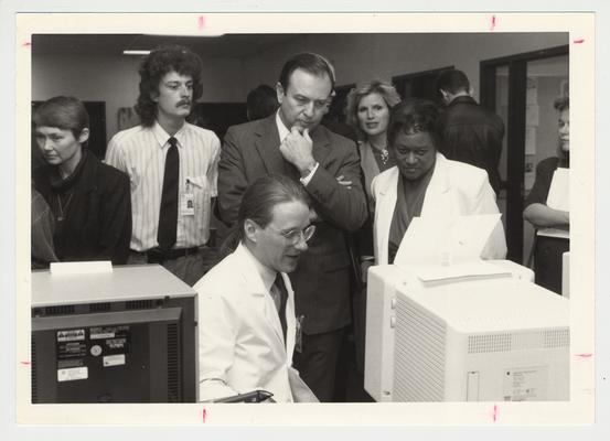President Wethington with Bradley Bryan (seated), second year medical student; Juanita Fleming (right of Wethington), Dean of the College of Nursing; and Janet Stith (far left), Director of the Medical Library.  There are also several unidentified people.  Everyone is looking at a computer in the Pathology Department.  Communi-K