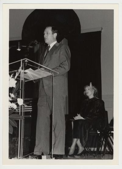 President Wethington is speaking at a podium.  Peggy Meszaros sits behind him in a cap and gown