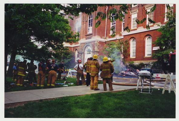 Unidentified fire fighters are standing outside of the Administration / Main Building  during the fire.  There is smoking debris on the ground in front of the fire fighters